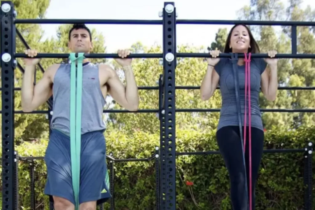 Casal fazendo barra fixa com auxílio de um super band. Casal treinando barra fixa ao ar livre.