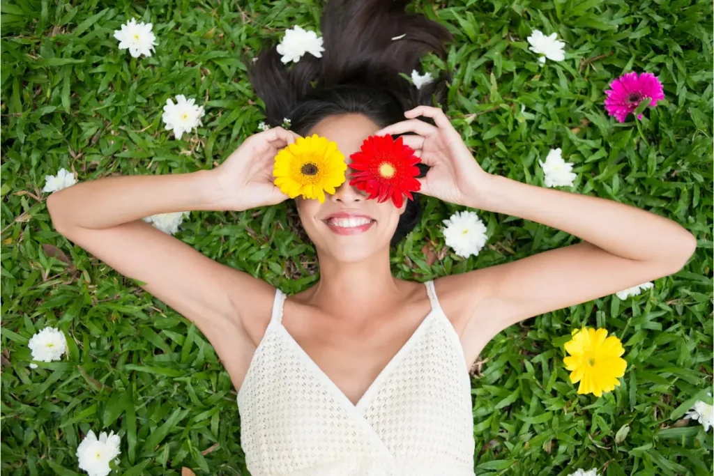 mulher sorridente deitada na grama verde com flores cobrindo os olhos, simbolizando felicidade e autoestima saudável ao ar livre